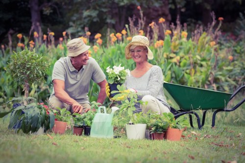 A mature couple is sitting in a lush, well-maintained backyard garden during daytime, surrounded by a variety of potted plants and garden tools. The man, wearing a straw hat and casual grey T-shirt, is engaging in planting or tending to the greenery. The woman, also wearing a straw sunhat and light-coloured clothing, is smiling and holding a flowering plant in a white pot, suggesting they are involved in gardening activities. Behind them, a flower bed with purple and orange blossoms is visible, alongside tall green shrubs and small trees that create a natural boundary for the outdoor space. To the right, there is a dark green wheelbarrow partially visible, hinting at ongoing garden work. The scene is bathed in natural daylight with a slightly cloudy sky, creating soft lighting that underscores the relaxing and productive atmosphere of outdoor garden maintenance, reflecting professional landscaping and gardening efforts typical of Southgate outdoor spaces.