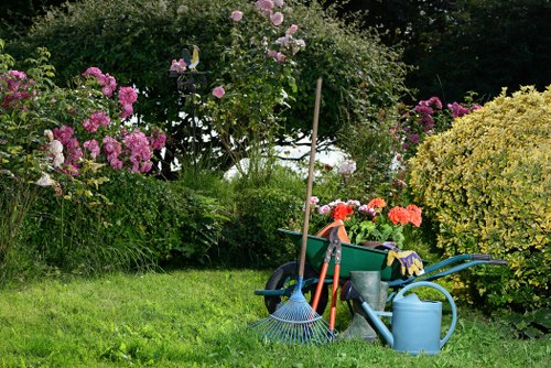 The image depicts a well-maintained garden area featuring a lush, green lawn in the foreground, with tools including a rake, a garden fork, and a watering can placed on the grass, suggesting ongoing gardening activities. In the background, there is a variety of flowering bushes and shrubs, including pink and purple blossoms, arranged along the edge of the garden, creating a vibrant and natural backdrop. A small tree with light pink flowers is also present, adding vertical interest to the scene. The garden is partly shaded by taller trees, with dappled sunlight filtering through the foliage, indicating a clear, possibly sunny day. The area appears to be a landscaped backyard or front garden, showcasing a tidy and inviting outdoor space suitable for gardening services such as lawn care, hedge trimming, and planting. The presence of gardening tools and watering equipment emphasizes the active maintenance and care of the garden environment, reflecting attention to garden aesthetics and healthy plant growth associated with professional landscaping efforts in Southgate or surrounding areas.