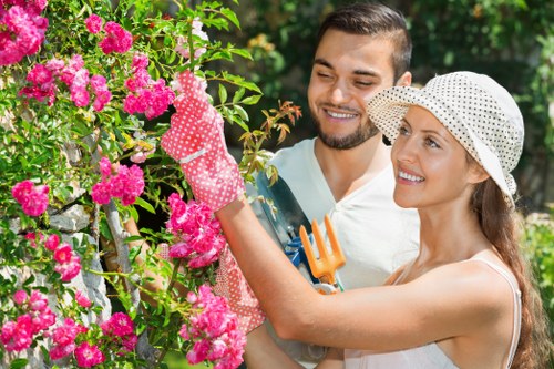 A young man and woman engaging in gardening in a lush, well-maintained garden with vibrant pink flowering bushes. The man, with short dark hair and a beard, is smiling and observing the woman's activities, while the woman, wearing a white sun hat and a light sleeveless top, is smiling and using pink gardening gloves to prune or tend to the flowering shrub. The garden features a mix of dense green foliage, flowering plants, and possibly other shrubs, suggesting a neatly landscaped outdoor space typical of residential gardens in Southgate. Natural daylight illuminates the scene, highlighting the vibrant colors of the flowers and foliage, with a blurred green background indicating a lush, inviting outdoor setting suitable for gardening and landscaping projects. This image captures the enjoyment of outdoor gardening, a key aspect of gardening services offered by companies such as Landscapers Southgate in the local area.