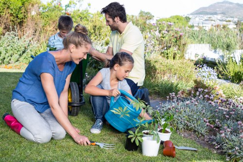A family gardening in a well-maintained backyard garden, with a lush green lawn area in the foreground, surrounded by flower beds filled with colorful plants and flowering shrubs, and bordered by a low hedge. The mother is kneeling on the grass, planting or tending to a young plant in the soil, while the daughter, sitting on the lawn, is handling watering cans and garden tools; the father and son are in the background, standing and engaging in gardening activities. The scene shows a bright, sunny day with clear skies, natural light illuminating the outdoor space, which features a mix of grass, soil, and decorative garden elements, creating a peaceful and inviting environment that highlights outdoor gardening and landscaping activities suitable for homes in Southgate or similar suburban areas.