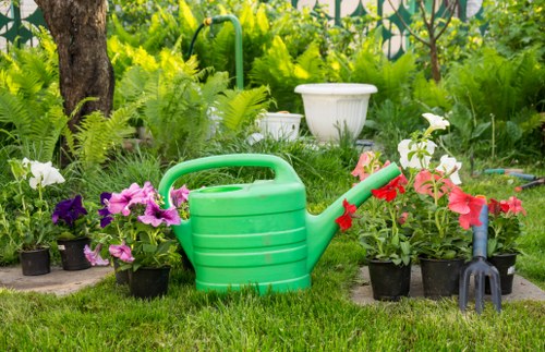 A garden scene featuring a variety of potted flowering plants, including pink and white blossoms, arranged along a well-maintained grassy lawn. A green plastic watering can with a curved handle and a red spout is prominently placed in the foreground, surrounded by the potted plants. Behind the plants, there are natural elements such as a tree trunk and lush green foliage, with additional garden features like a white decorative planter and a garden rake with a black handle visible. The outdoor space appears to be part of a landscaped yard in Southgate, with even, vibrant grass, and the scene is lit by natural daylight, suggesting a bright, clear day. The arrangement and elements hint at gardening activities and landscape maintenance, with a focus on nurturing plants and maintaining the garden's aesthetic appeal.