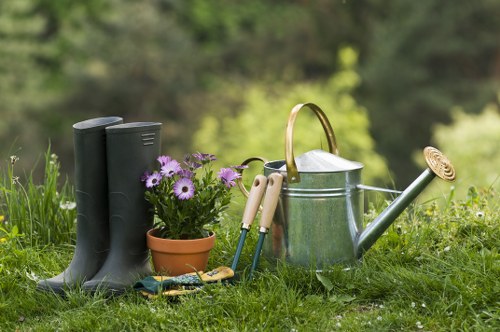 On a lush green lawn in an outdoor garden setting, there are a pair of black rubber gardening boots standing upright next to a small terracotta pot filled with purple daisy-like flowers. Nearby, a collection of garden tools with light wooden handles, including a hand trowel and a rake, are laid on the grass. A silver metal watering can with a long spout and wooden handle is positioned beside the tools, leaning slightly inward. The background features blurred trees and foliage, indicating a natural, well-maintained outdoor space typical of a backyard or landscaped area in Southgate. The scene is well-lit with natural daylight, suggesting a clear weather day, and the overall environment reflects a tidy, cultivated garden prepared for ongoing planting or watering activities, consistent with professional gardening and landscaping services offered by Landscapers Southgate.