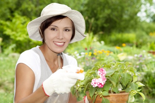 The image shows a woman in a garden during daytime, wearing a wide-brimmed sun hat, a white short-sleeved shirt, and gardening gloves, holding a terracotta pot filled with pink and white flowering plants. In the background, there are lush green trees, colourful flower beds, and a well-maintained lawn area, indicating a landscaped outdoor space typical of Southgate gardens. She appears to be engaged in planting or tending to the garden, with a gentle smile on her face, suggesting enjoyment of outdoor gardening activities. The garden features a mix of flowering shrubs, grass, and possibly decorative borders, all aligned with professional garden renovation and landscaping services offered by Landscapers Southgate. The lighting is natural, indicating a clear day, with vibrant colours and crisp details emphasizing the healthy foliage and floral elements, characteristic of well-kept residential gardens in the Southgate area. This scene illustrates outdoor gardening work suitable for enhancing or renovating garden spaces in the local community with professional landscaping expertise.
