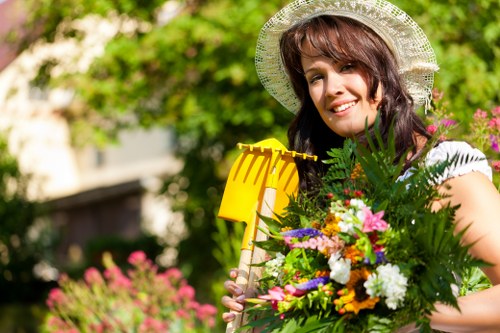 A woman wearing a straw sunhat and a white top stands in a lush garden, holding a yellow garden tool in one hand and a large, colorful bouquet of various flowers and greenery in the other. The garden features a well-maintained lawn with dense, green grass, bordered by flower beds filled with blooming flowers and shrubs. In the background, there are mature trees with green foliage providing partial shade, and a blurred garden structure or fence. The scene appears to be outdoors on a sunny day, with natural light illuminating the vibrant plant life and creating a bright, cheerful atmosphere consistent with landscape gardening and outdoor maintenance services in Southgate or nearby areas.