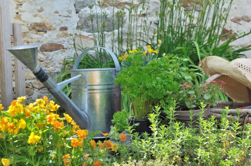 The image depicts a small, well-maintained garden area featuring a variety of plants and garden tools. In the foreground, there are vibrant yellow and orange flowering plants with lush green foliage, creating a lively contrast. Behind these, a black plastic container holds a bushy green plant with delicate leaves, while a silver metal watering can with a curved handle sits nearby, suggesting ongoing garden care. To the right, a beige straw hat rests on a dark-colored potted plant, possibly on a garden bench or ledge. The background includes tall, slender green reeds or grasses growing along a stone wall, providing a natural backdrop to the garden scene. The overall environment appears to be outdoors, with natural lighting indicating a bright, sunny day, and the setting emphasizes outdoor gardening activities, typical of an outdoor space maintained by gardening services such as those offered by landscaperssouthgate.co.uk in Southgate, London, Gunoard. The setup reflects a thriving garden space ready for ongoing planting and maintenance activities.