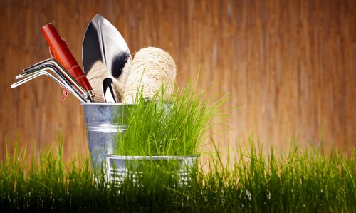 A close-up of a gardening collection featuring a small metal bucket containing various hand tools such as a trowel, a cultivator, and a rake, along with a beige ball of garden twine, all situated amidst lush, green grass outdoors. The tools have wooden and metal handles, and the background showcases a warm, natural wooden surface. The scene emphasizes garden maintenance and outdoor skills, aligning with landscaping and gardening services offered by Landscapers Southgate, set within a well-maintained garden or outdoor space with natural lighting that highlights the vibrant greenery and the tool details.