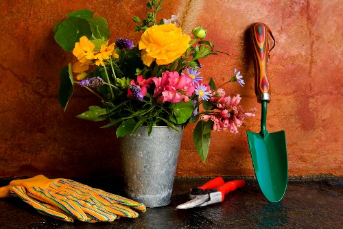 A close-up image showing a concrete indoor surface with a large pot of freshly cut, colorful flowers including yellow, pink, purple, and blue blooms, arranged in a natural, slightly wild style. The flowers sit on a dark workspace with gardening tools nearby, including a small green trowel with a red handle, a pair of pruning shears, and a pair of yellow and green striped gardening gloves, suggesting preparation for outdoor gardening or landscaping tasks. The background features a warm-toned wall, indicating an indoor environment used for plant care or garden planning, in a setting that reflects professional gardening or landscaping activity in Southgate, London, with a focus on plant arrangement and outdoor maintenance.