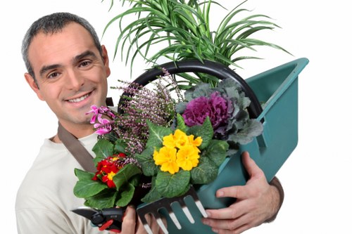A smiling man with short dark hair, wearing a light-colored shirt, holding a green plastic garden trowel and a small gardening basket filled with colorful flowering plants and foliage. The basket contains yellow, purple, pink, and red flowering plants with green leaves, set against a plain, light background. The scene captures an outdoor gardening or landscaping activity, highlighting plant cultivation and garden care, relevant to services offered by Landscapers Southgate near the Southgate area. The garden elements suggest a well-maintained, vibrant outdoor space suitable for gardening and landscaping improvements.