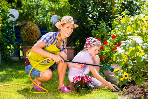 In a lush backyard garden with vibrant green foliage and colorful flowering plants, a woman and a young girl are engaged in gardening activities. The woman, dressed in a yellow apron, plaid shirt, and straw hat, is kneeling on the grass and holding a garden hose, smiling as she looks towards the camera. The girl, wearing a floral hat and casual clothing, is focused on planting or watering flowers in a flower bed bordered by a wooden edge. The garden features a well-maintained lawn with dense bushes and small trees in the background, illuminated by natural sunlight, creating a bright and inviting outdoor space. Decorative garden lights are visible among the plants, adding to the organized and cared-for landscape, which is typical of residential gardens in Southgate. This scene highlights outdoor gardening activities suitable for a professional landscaper’s range of services, including planting, lawn care, and garden maintenance, ideal for enhancing garden beauty and functionality. The setting appears to be a well-kept private garden, emphasizing natural tones and an inviting atmosphere in a suburban environment.