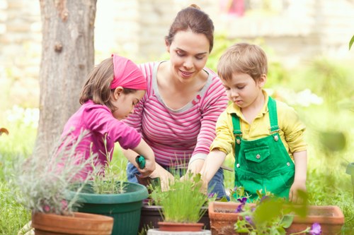 A woman, likely a gardener or parent, is shown in a backyard garden with two young children; she is wearing a pink and white striped top, and the children are dressed in colorful outdoor clothing with one in a pink headscarf and the other in green overalls. They are gathered around a collection of garden pots, some filled with blooming flowers and others with small green plants or seedlings, positioned on a wooden surface or garden table. The garden features a large tree trunk in the background, indicating mature landscaping, with neatly tended grass, flowering plants, and shrubs creating a lush outdoor environment. The scene is illuminated by natural daylight, suggesting a mild, pleasant weather typical of Southgate or nearby areas, reinforcing the connection to outdoor gardening and landscape maintenance services provided by Landscapers Southgate. This image visually captures the joy and engagement of outdoor gardening activities, reflecting the importance of well-maintained, aesthetically pleasing garden spaces in residential properties across the local area.