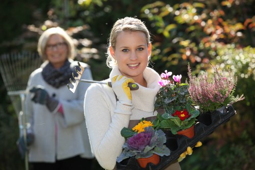 A young woman in a white sweater holding a black tray with a variety of colorful potted flowering plants, including purple, yellow, and red blooms, in a garden setting during daytime. Behind her, an older woman with glasses, a white jacket, and a scarf is smiling and holding garden tools, standing in a lush outdoor space with green foliage, hedges, and flowering shrubs. The garden has a well-maintained lawn with dense, healthy grass, bordered by flower beds and shrubs, brightly lit by natural sunlight, suggesting a clear day. The scene captures outdoor gardening activity typical of a landscaped garden in Southgate, with trees and plants providing natural tones and texture. The women are dressed appropriately for gardening, emphasizing the outdoor maintenance aspect of landscaping services provided by Landscapers Southgate.