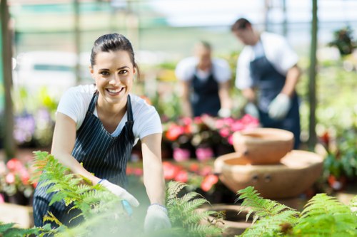 A smiling young woman wearing a striped apron and gardening gloves is tending to green ferns in a bright greenhouse or garden center, with colorful flowering plants and potted greenery arranged in the background. The scene includes a variety of garden elements such as lush grass, flowering shrubs, and a stone water fountain, suggesting a well-maintained outdoor space that could be part of a landscaping or gardening project in Southgate. Natural daylight filters through glass panels, illuminating the vibrant plant textures and earthy tones of the garden environment. This setting showcases professional gardening work aligned with services offered by landscapers Southgate, emphasizing attention to plant health, lawn care, and garden design, subtly supporting local SEO through the setting and visual context of garden maintenance and landscaping in the Southgate area.